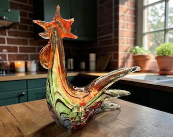May include: A colorful glass rooster sculpture with orange, green, and red hues. The decorative figurine has a detailed comb and tail, and is displayed on a wooden surface. The rooster is a decorative item.