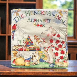 May include: A children's alphabet book titled "The Hungry Animal Alphabet Book." The cover features a hippo in a floral dress, a parrot, and various food items. The book is on a wooden surface.