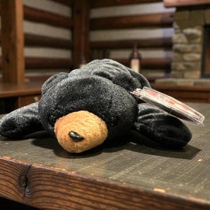 Può includere: Un orsacchiotto nero con muso marrone e naso nero, appoggiato su una superficie di legno scuro. L'orso ha un'etichetta attaccata. Lo sfondo comprende un camino e mobili in legno.