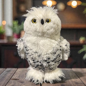 Pode incluir: Uma coruja de pelúcia, predominantemente branca com manchas pretas nas asas e nas costas. Tem olhos amarelos brilhantes e um bico preto. A coruja está em pé sobre uma superfície de madeira. O brinquedo tem uma textura macia e uma aparência amigável.