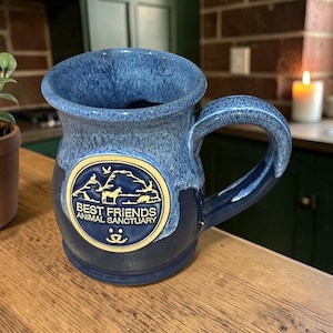 May include: A blue ceramic mug with a handle, featuring a gold emblem that reads "Best Friends Animal Sanctuary." The mug has a textured, glazed finish with darker blue at the base and lighter blue at the rim. It sits on a wooden surface.