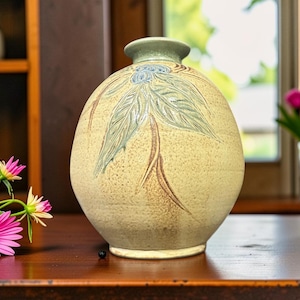 Peut inclure: Un vase en céramique rond avec une finition crème mouchetée, présentant un motif botanique sculpté de baies bleues et de feuilles vertes. Le vase possède un col court évasé et un corps arrondi lisse, offrant un aspect texturé et naturel.