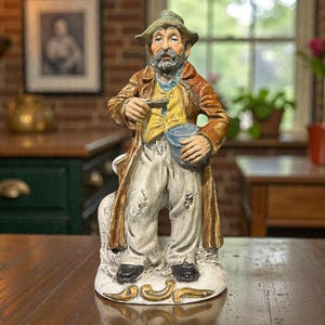 May include: A detailed ceramic figurine of an elderly man in tattered clothing, holding a small bucket and a tool. The figure wears a hat, long coat, and yellow vest. The base has gold accents.