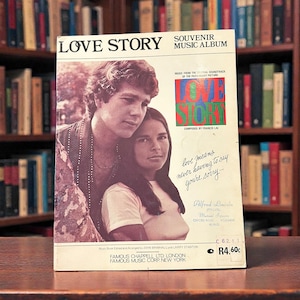 以下が含まれることがあります： カップルの写真が特徴の、ヴィンテージ「ラブ・ストーリー」の記念音楽アルバムのカバー。カバーには大きな文字でタイトルが書かれており、「love means never having to say you're sorry」というテキストがあります。背景には本棚が写っています。