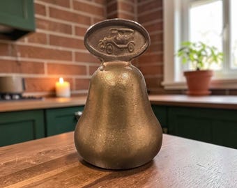 May include: A brass-coloured bell with a smooth, pear-shaped body and a flat, oval-shaped handle at the top. The handle features a raised, embossed relief design of a vintage automobile, showing the side profile of the car in a metallic finish.