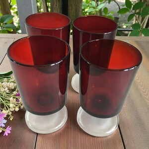 May include: Four red-tinted glass tumblers with a rounded shape, each resting on a white base. The glasses have a deep red colour and are arranged on a wooden surface. The image showcases a set of drinking glasses.