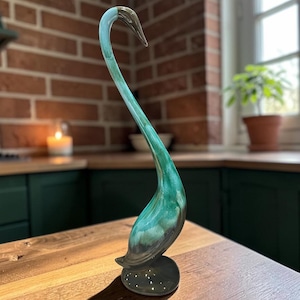 Può includere: Un'elegante statuetta a forma di cigno in ceramica con un lungo collo curvo. Il cigno presenta una sfumatura di tonalità verde acqua e verde scuro, con una base scura. La scultura è posizionata su una superficie di legno.