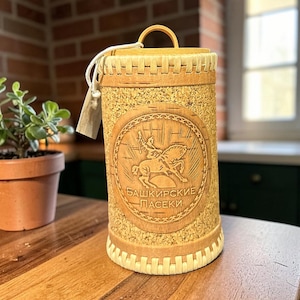 May include: A cylindrical, light brown birch bark container with a handle and a decorative carved emblem. The emblem features a person on horseback. The container is on a wooden surface.