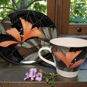May include: A black and white floral teacup and saucer set. The teacup and saucer feature a large orange flower design. The teacup has a white interior and handle. The saucer is round. A small bouquet of purple flowers sits in front of the teacup.