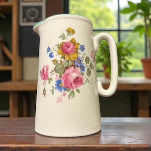 May include: Cream-colored ceramic pitcher with a large handle and a floral design. The flowers are pink, yellow, and blue, with green leaves. The rim of the pitcher is trimmed with green.