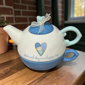 May include: A white and blue ceramic teapot with a matching teacup base. The teapot features a blue heart design with the word "love" and the phrase "Start each day in a happy way." The teapot has a blue handle and lid.