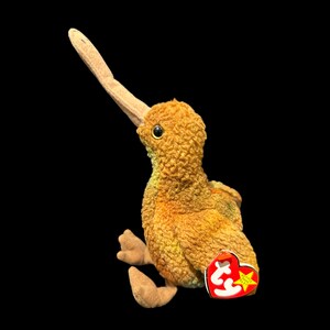 May include: A brown and yellow stuffed kiwi bird with a long orange beak. The bird is sitting on a black background and has a red and yellow Beanie Baby tag attached to its leg.