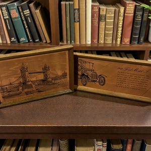 May include: Two framed wooden plaques with etched designs. One depicts the Tower Bridge in London, the other a vintage Ford Model T car with text. The frames are a light brown color.