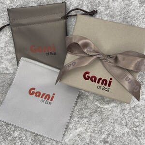 Op de afbeelding: Twee cadeaudozen in bruin en wit. Beide dozen hebben een rood logo met de tekst "Garni of Bali". Een doos is een klein zakje met een trekkoord. De andere doos is een grotere doos met een bruin satijnen lint eromheen gebonden.