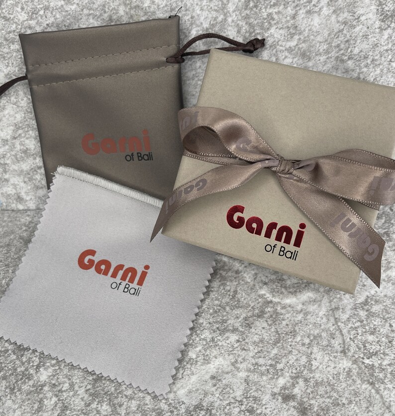 Op de afbeelding: Twee cadeaudozen in bruin en wit. Beide dozen hebben een rood logo met de tekst "Garni of Bali". Een doos is een klein zakje met een trekkoord. De andere doos is een grotere doos met een bruin satijnen lint eromheen gebonden.