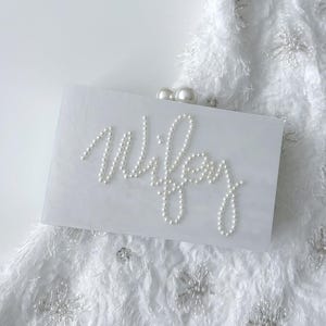 Peut inclure: Une pochette rectangulaire blanche avec le mot "Wifey" &eacute;pel&eacute; en perles blanches. La pochette a un fermoir en perle et repose sur un tissu blanc textur&eacute; avec des embellissements en forme de flocons de neige.