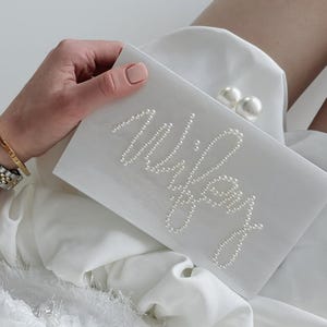 Peut inclure: Une pochette blanche avec le mot "Wifey" &eacute;pel&eacute; en petites perles. La pochette a un fermoir en perles. La personne tenant le sac porte un bracelet dor&eacute; et une montre.