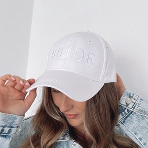May include: White baseball cap with the word "BRIDE" embroidered on the front. The cap is worn by a person wearing a light blue denim jacket with pearl embellishments.