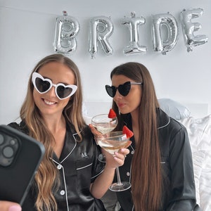 May include: Two people wearing black satin pyjamas with white piping and heart-shaped sunglasses are holding champagne flutes and taking a selfie. The word "BRIDE" is spelled out in silver balloons behind them.