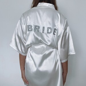 Puede incluir: Bata de satén blanco con mangas cortas y cinturón. La parte posterior de la bata tiene la palabra "BRIDE" escrita en purpurina plateada.