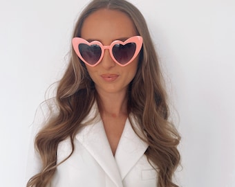 May include: A woman wearing a white blazer with a heart-shaped pink sunglasses.