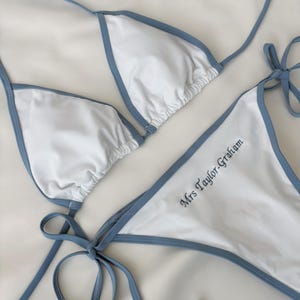 May include: White bikini set with light blue trim and ties. The bikini top is a triangle style, and the bottoms feature the embroidered text "Mrs Taylor-Graham". The swimwear is displayed on a white surface.