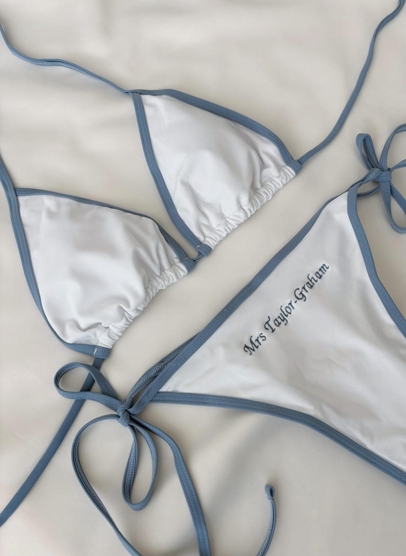 May include: White bikini set with light blue trim and ties. The bikini top is a triangle style, and the bottoms feature the embroidered text "Mrs Taylor-Graham". The swimwear is displayed on a white surface.