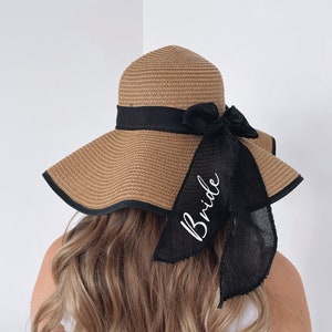 May include: A brown straw sun hat with a black ribbon and bow. The ribbon has the word "Bride" written in white script.