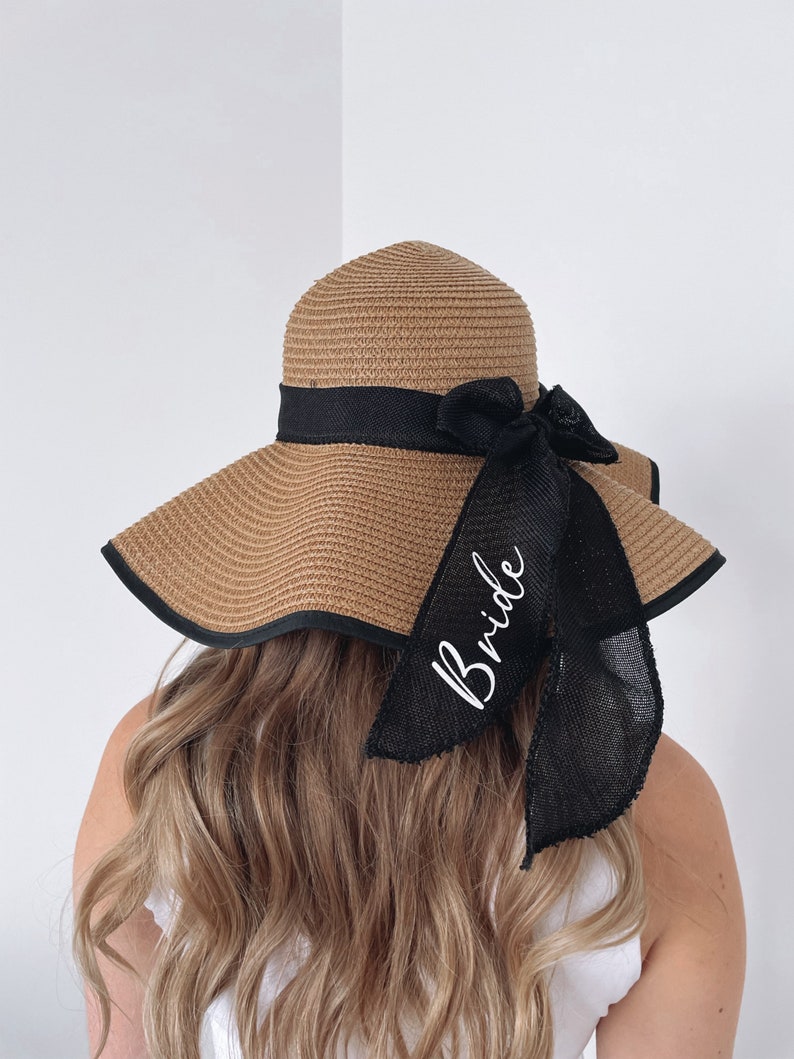 May include: A brown straw sun hat with a black ribbon and bow. The ribbon has the word "Bride" written in white script.
