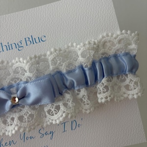 May include: A light blue satin and lace wedding garter with a small bow and a rhinestone accent. The garter is displayed on a white card with the text "Something Blue" and "When You Say 'I Do'" in blue script.