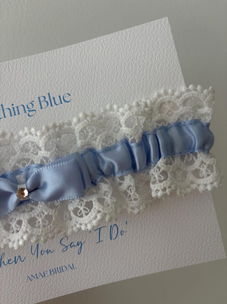 May include: A light blue satin and lace wedding garter with a small bow and a rhinestone accent. The garter is displayed on a white card with the text "Something Blue" and "When You Say 'I Do'" in blue script.