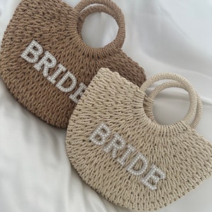 May include: Two straw tote bags with a pearl embellished "BRIDE" design. One bag is brown and the other is beige.