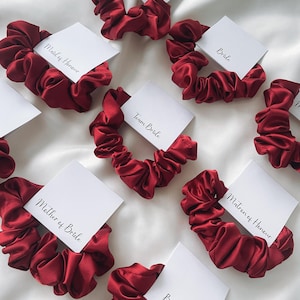 Könnte beinhalten: Burgunderrote Satin-Haargummis mit weißen Karten, auf denen "Bride", "Bridesmaid", "Maid of Honor", "Matron of Honor", "Mother of Bride" und "Team Bride" steht.