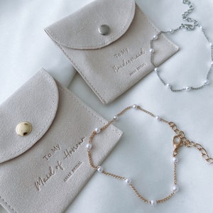 May include: Two bracelets with faux pearls and matching pouches. One bracelet is silver-toned, the other gold-toned. The pouches are beige with the text "To My Bridesmaid" and "To My Maid of Honour" in gold lettering. The brand name "AMAE BRIDAL" is also visible.