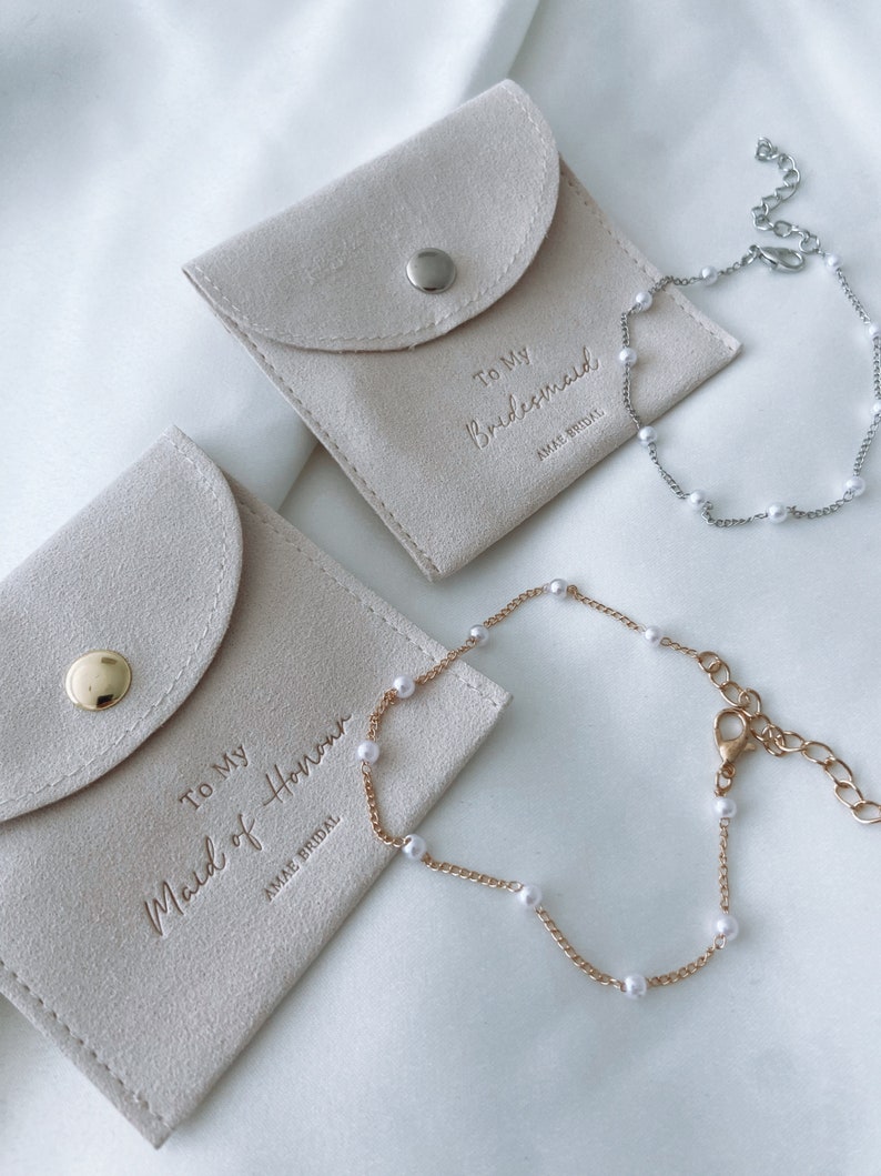 May include: Two bracelets with faux pearls and matching pouches. One bracelet is silver-toned, the other gold-toned. The pouches are beige with the text "To My Bridesmaid" and "To My Maid of Honour" in gold lettering. The brand name "AMAE BRIDAL" is also visible.