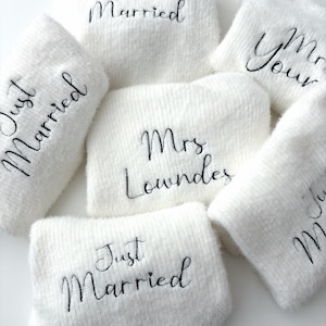Può includere: Un gruppo di asciugamani bianchi e soffici con ricami neri. Le scritte includono "Just Married", "Mrs. Lowndes" e "Mr & Mrs Your". Gli asciugamani sono disposti in modo leggermente sovrapposto, adatti per un matrimonio.