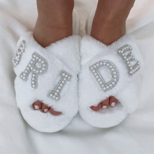 May include: White fluffy slippers with the word "BRIDE" spelled out in pearl-studded letters. The slippers are on a white surface.