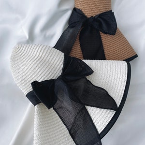 May include: Two straw sun hats with wide brims and black ribbon bows. The hats are in different colors, one is white and the other is brown.
