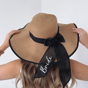 May include: A brown straw sun hat with a black ribbon and bow. The ribbon has the word "Bride" embroidered on it in white.