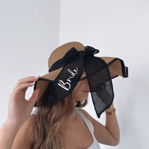 May include: A brown straw sun hat with a black ribbon and a white "Bride" inscription. The ribbon is tied in a bow at the back of the hat.
