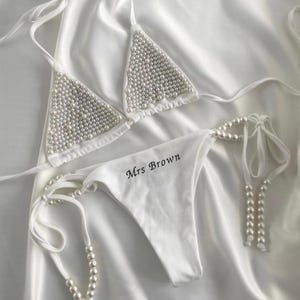 May include: White bikini set with pearl embellishments. The triangle top and the bottom are adorned with numerous small pearls. The bottom of the bikini has the text "Mrs Brown" printed on it. The set is displayed on a white satin fabric.