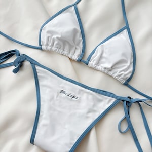 May include: White bikini set with blue trim. The top is a triangle style with adjustable straps. The bottom features the embroidered name "Mrs Lopez". The bikini is made of a smooth, likely synthetic fabric.