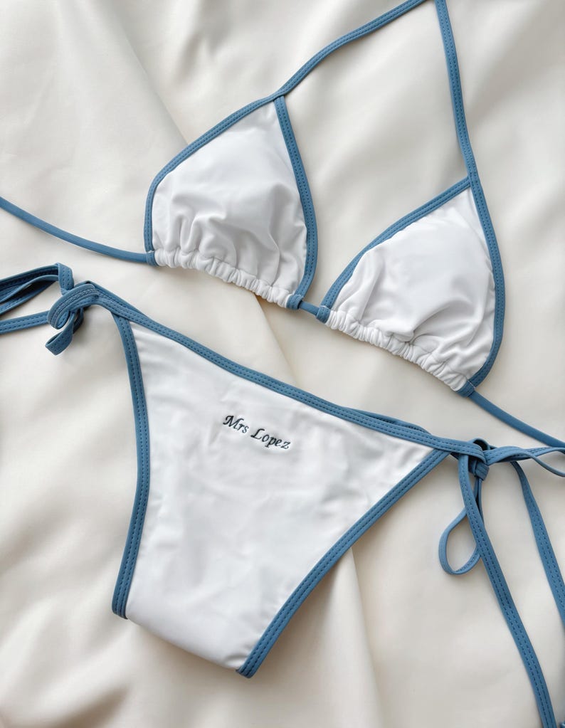 May include: White bikini set with blue trim. The top is a triangle style with adjustable straps. The bottom features the embroidered name "Mrs Lopez". The bikini is made of a smooth, likely synthetic fabric.