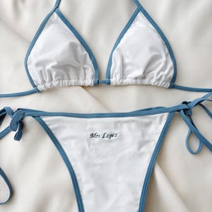 May include: White bikini set with blue trim. The top is a triangle style with halter straps. The bottom features side ties and the embroidered text "Mrs Lopez".