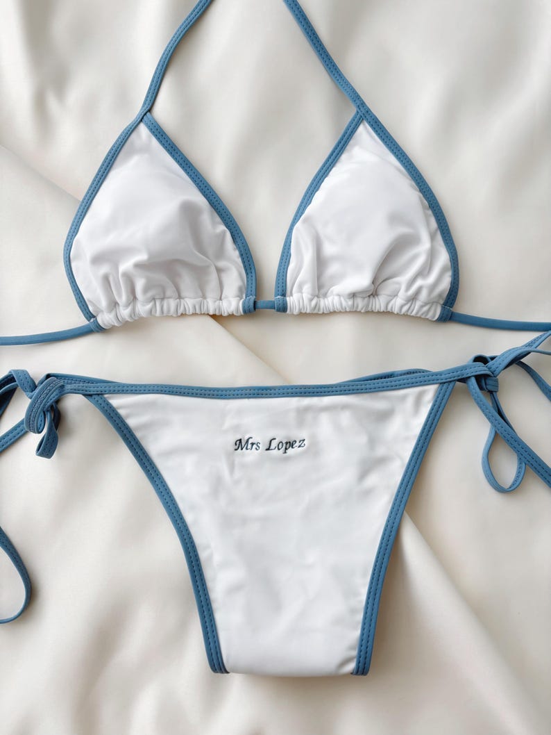 May include: White bikini set with blue trim. The top is a triangle style with halter straps. The bottom features side ties and the embroidered text "Mrs Lopez".