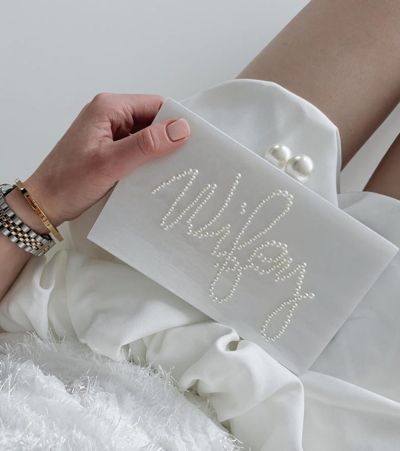 Peut inclure: Une pochette blanche avec le mot "Wifey" &eacute;pel&eacute; en petites perles. La pochette a un fermoir en perles. La personne tenant le sac porte un bracelet dor&eacute; et une montre.
