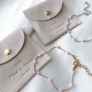 May include: Two light beige suede pouches with the text "To My Bridesmaid" and "To My Maid of Honor" embossed on them. Each pouch has a matching pearl bracelet. One bracelet is gold-toned, the other silver-toned, both with white pearls.