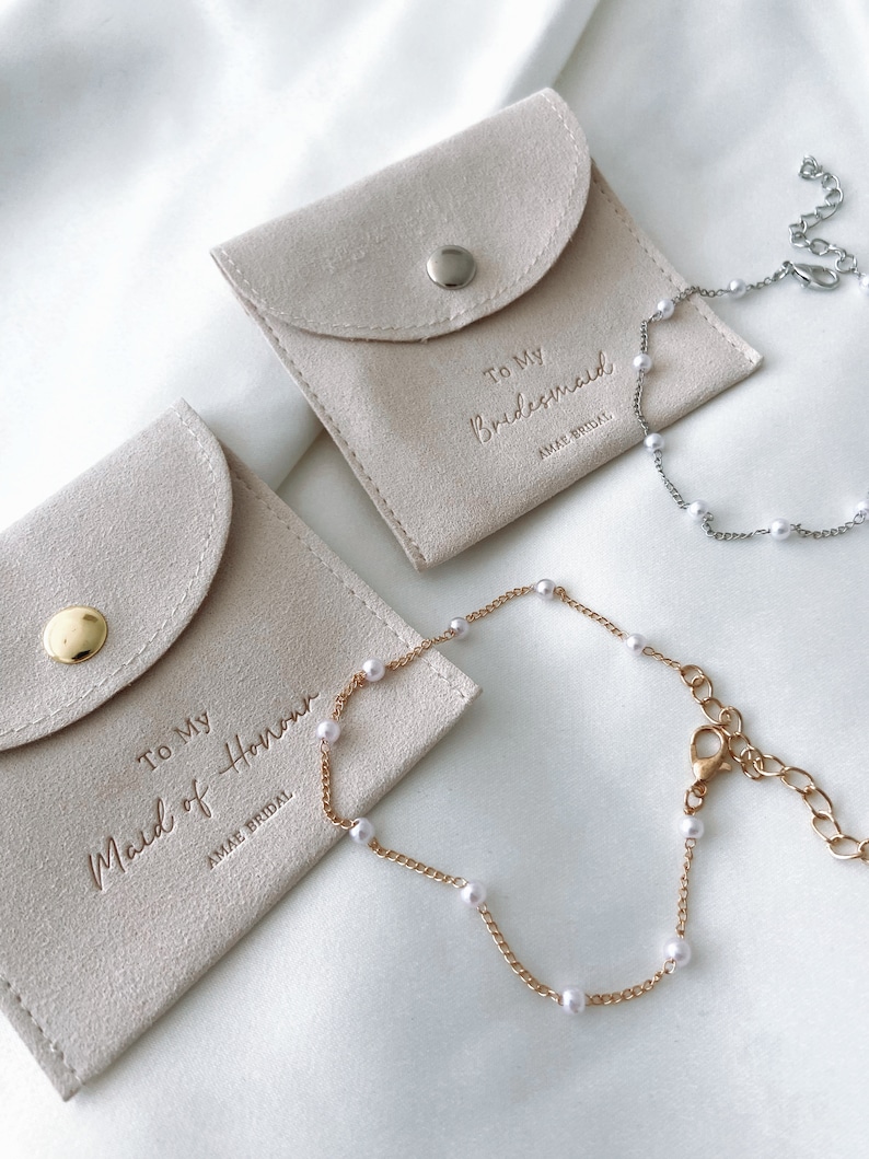 May include: Two light beige suede pouches with the text "To My Bridesmaid" and "To My Maid of Honor" embossed on them. Each pouch has a matching pearl bracelet. One bracelet is gold-toned, the other silver-toned, both with white pearls.