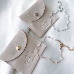 May include: Two light beige suede pouches with the text "To My Bridesmaid" and "To My Maid of Honor" embossed on them. A silver and a gold bracelet with small white pearls are next to the pouches. The pouches have a snap closure.