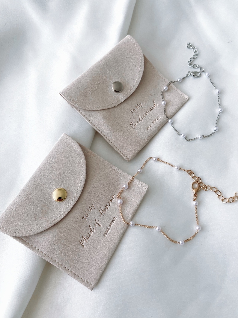 May include: Two light beige suede pouches with the text "To My Bridesmaid" and "To My Maid of Honor" embossed on them. A silver and a gold bracelet with small white pearls are next to the pouches. The pouches have a snap closure.